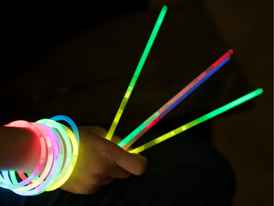 A hand holding glow in the dark sticks. The wrist of the person's arm is wearing many glow in the dark bracelets.