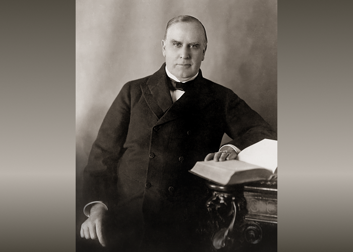 President William McKinley sitting at desk with hand resting on an open book.
